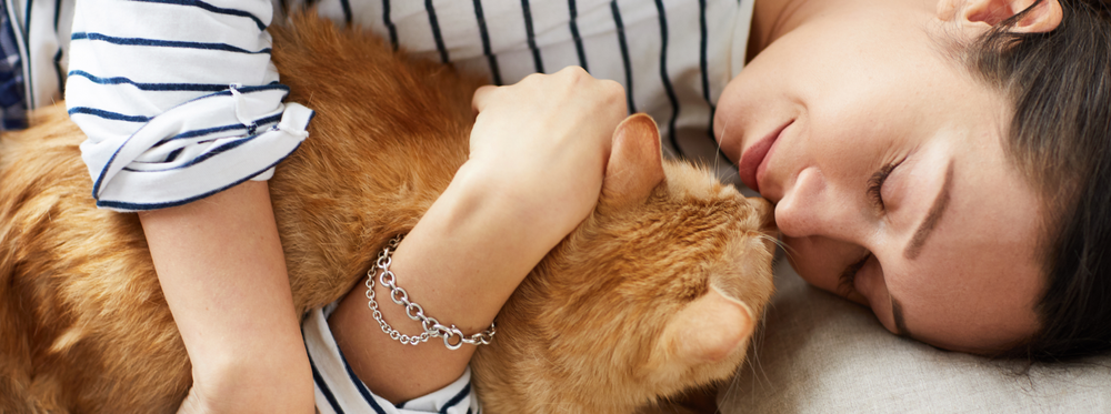 Femme allongée, yeux fermés, câlinant un chat roux en souriant.